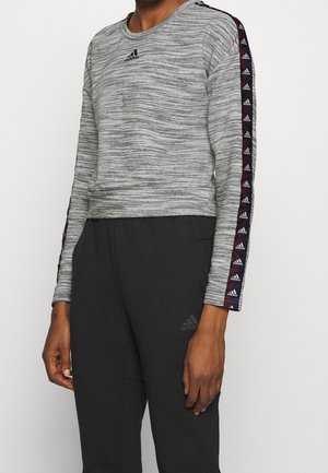 Persoon draagt een grijs Adidas shirt met lange mouwen, zwart en paarse logostrepen op de mouwen, en zwarte Adidas broek.