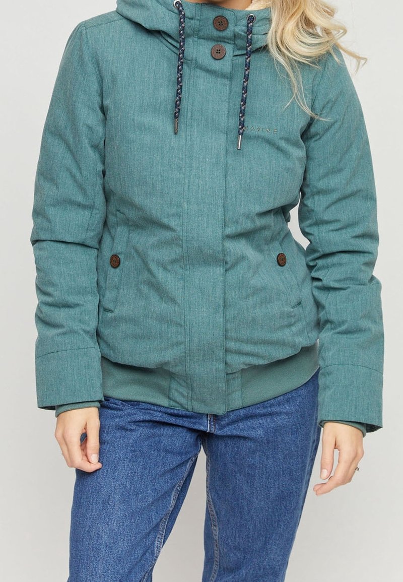 Mujer con chaqueta de invierno con capucha de color verde azulado con botones marrones y cordones, combinada con jeans de mezclilla azul, sobre un fondo liso.