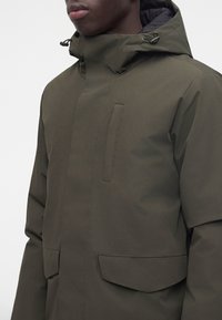 Homme portant un manteau d'hiver vert olive foncé avec capuche, col montant et trois poches visibles, visible du torse jusqu'au milieu du visage.