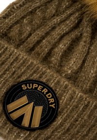 Cappello di lana marrone lavorato a maglia con texture a costine, caratterizzato da una patch circolare nera con "Superdry" in oro e un design geometrico.