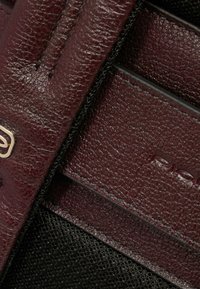 Portafoglio in pelle bordeaux con superficie testurizzata, caratterizzato da dettagli in tessuto nero e logo dorato. Design compatto e rettangolare.