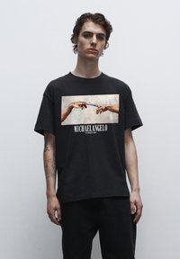 Sort bomulds t-shirt med et frontprint af to hænder, der strækker sig mod hinanden, med teksten "MICHAELANGELO" nedenfor billedet.