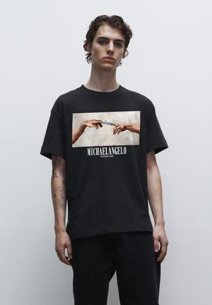 Schwarzes Baumwoll-T-Shirt mit einem Frontdruck von zwei Händen, die sich entgegenstrecken, und dem Text "MICHAELANGELO" unter dem Bild.