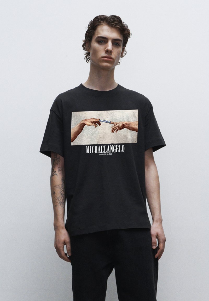 Sort bomulds t-shirt med et frontprint af to hænder, der strækker sig mod hinanden, med teksten "MICHAELANGELO" nedenfor billedet.