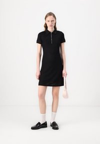 Fekete, rövid ujjú pólóruha cipzárral elöl, textúrált anyagból, slim fit, fekete fűzős cipővel és fehér zoknival párosítva. Kis rózsaszín táska.