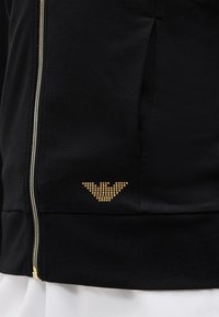 Čierna bunda so zlatým cvočkovým emblémom na dolnej strane, vybavená predným zipsom a bočným vreckom, nosená cez biely odev.