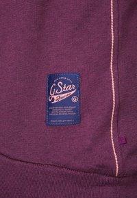Tessuto di felpa viola con un'etichetta testurizzata. L'etichetta presenta uno sfondo blu, testo bianco e rosa, ed è cucita con un filo chiaro sottile.