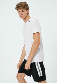 Hvid atletisk T-shirt med korte ærmer og et sort logo, kombineret med sorte shorts med en侧paneleffekt i hvid tekst.
