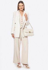 Donna che indossa un blazer bianco, un gilet beige con bottoni, pantaloni a gamba larga, sandali floreali e porta una borsa bianca con tracolla dorata.