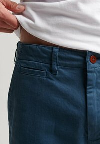 Pantalones azul oscuro con una textura suave, costuras visibles y un botón marrón. El detalle del bolsillo y la cinturilla son parcialmente visibles.