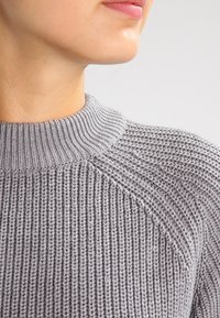 Grijze gebreide trui met een geribde halslijn en een textuurpatroon, met een close-up van de steken en stofdetails.