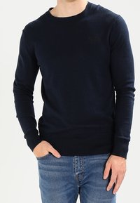 Tmavě modrý pletený svetr s dlouhými rukávy a kulatým výstřihem, zdobený nenápadným logem na hrudi a žebrovanými manžetami a lemem.