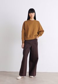 Donna con lunghi capelli neri che indossa una felpa oversize marrone, pantaloni palazzo marroni scuri e scarpe bianche, in piedi di fronte a una parete bianca semplice.