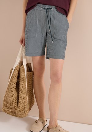 Person iført marineblå og hvide stribede shorts med snøre, beige slip-on sko, der holder en stor vævet taske med hvide håndtag.