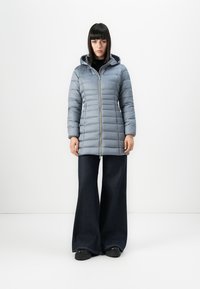 Lichtblauwe pufferjas met verticale stiksels, een capuchon en een rits aan de voorkant. Gedragen boven wijde, donkere denim broeken en zwarte schoenen.