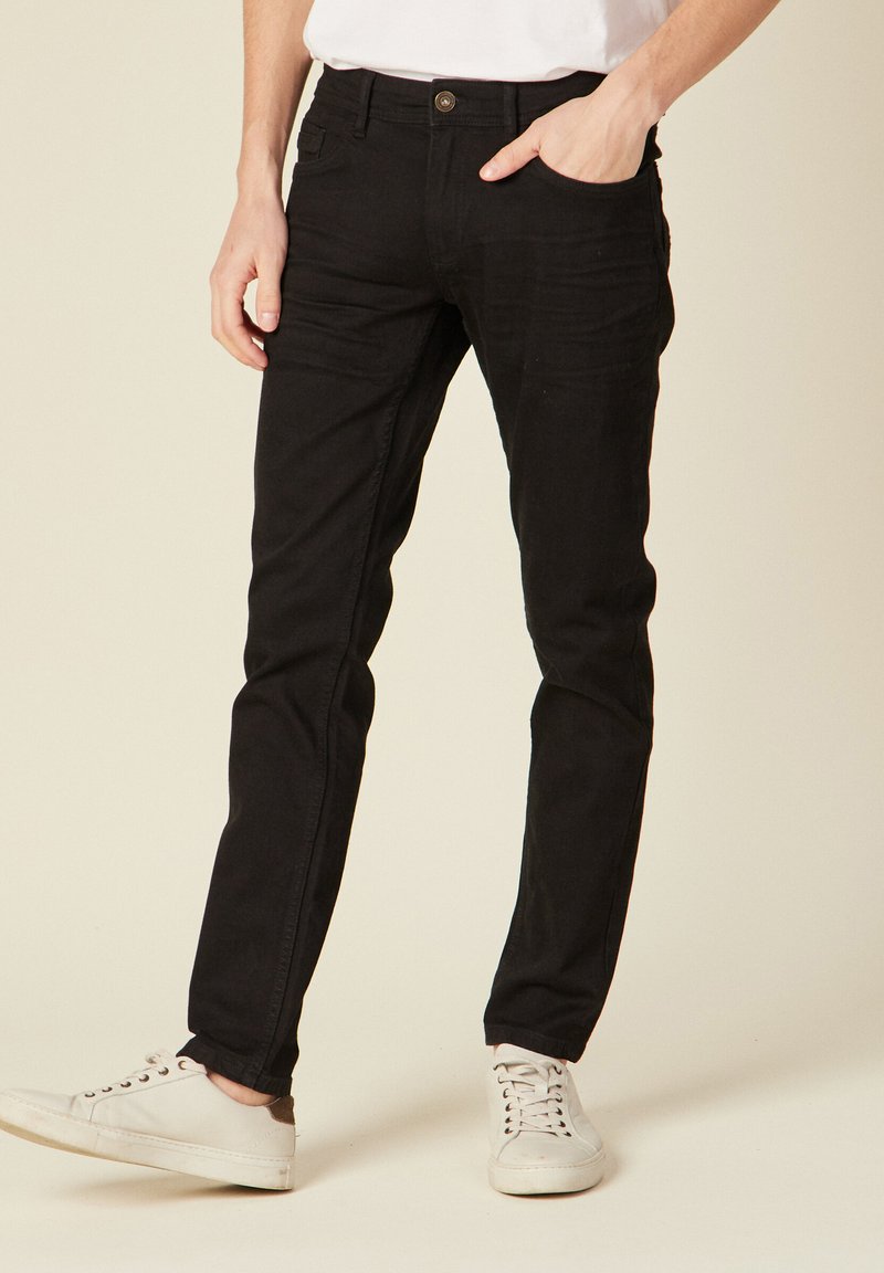 Schwarze Slim-Fit-Jeans aus weichem, strukturiertem Material mit Vorder- und Hintertaschen, getragen mit weißen Sneakers, ausgestattet mit einem klassischen Knopfverschluss.