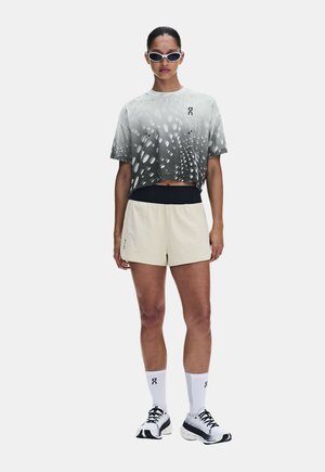 Kvinde står iført hvid og grå mønstret crop top, beige shorts, hvide træningsstrømper, hvide og sorte løbesko og hvide solbriller.