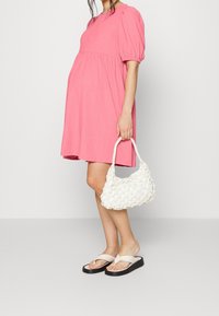 Donna in un vestito corto a maniche a sbuffo rosa, che tiene una borsa in fibra bianca, indossando ciabatte platform beige, in piedi su uno sfondo neutro.