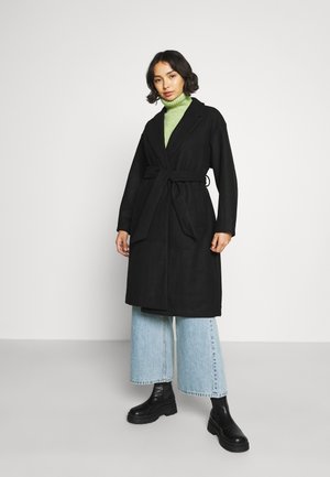 Black belted coat with a shawl collar, worn over a green turtleneck, paired with wide light blue jeans and chunky black boots.