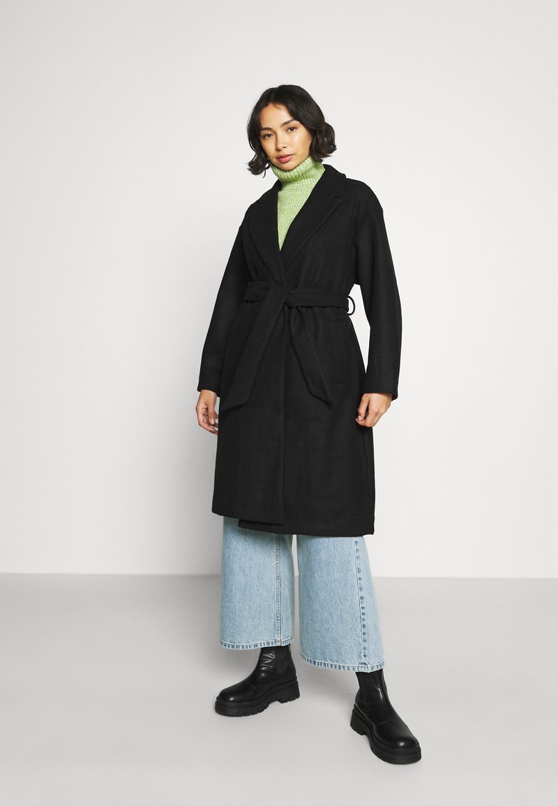 Black belted coat with a shawl collar, worn over a green turtleneck, paired with wide light blue jeans and chunky black boots.