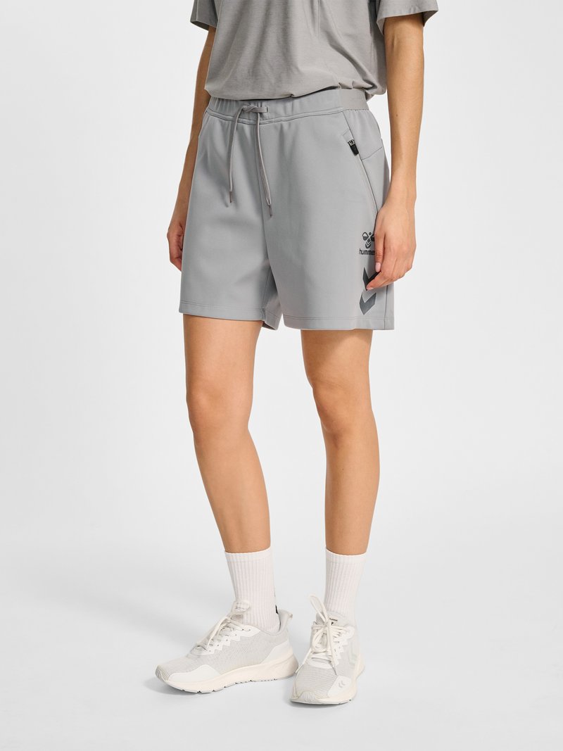 Personne portant un short gris clair avec cordon, un t-shirt ample assorti, des chaussettes blanches montantes et des baskets blanches, debout devant un fond uni.