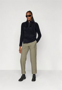 Zwarte fleece trui met een halve ritssluiting bij de kraag, gecombineerd met licht olijfgroene broek en zwarte schoenen. Model draagt een zonnebril.