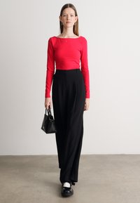 Rdeča bluzica z dolgimi rokavi, črne hlače visoko pasu, črna torbica in črne čevlje z belimi detajli, postavljeni na nevtralni ozadju.