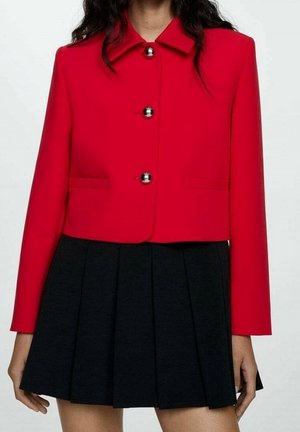 Žena oblečená v krátké červené bundě se stříbrnými knoflíky přes černou plisovanou mini sukni, zobrazena od krku po střed stehen.