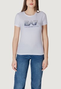 T-shirt bianco aderente con un logo grafico "EA7 Emporio Armani" in blu, maniche corte e design a girocollo, abbinato a jeans blu.