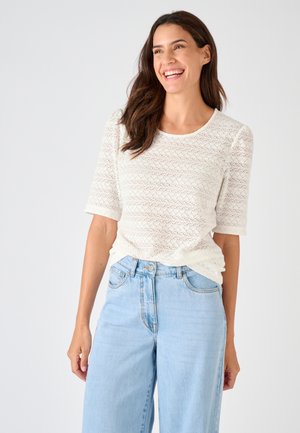 Lachende vrouw met lang bruin haar, die een witte getextureerde top met korte mouwen en lichtblauwe spijkerbroek met hoge taille draagt, tegen een effen achtergrond.