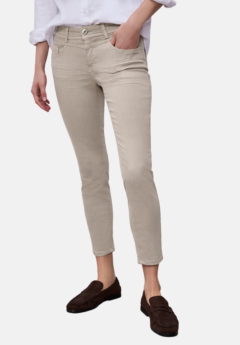 Beige, tætsiddende bukser båret med hvid skjorte, venstre hånd i lommen, kombineret med mørkebrune loafers på ensfarvet baggrund.