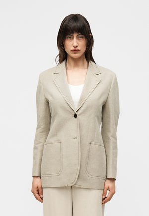 Žena s tmavými vlasmi, oblečená v béžovom saku s dvoma prednými vreckami cez biely top, stojaca pred jednoliatym svetlým pozadím.