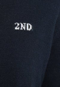 Tumši zils džemperis ar adījuma rakstu un baltu izšūtu uzrakstu "2ND" priekšpusē.