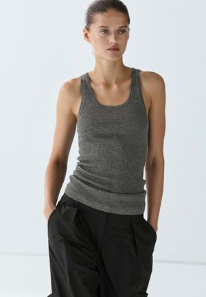 Femme aux cheveux attachés portant un débardeur gris côtelé et un pantalon noir ample, debout les mains dans les poches devant un fond uni.