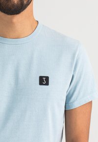 Hellblauses Baumwoll-T-Shirt mit rundem Halsausschnitt, ausgestattet mit einem kleinen schwarzen Quadrat-Patch mit dem weißen Buchstaben "J" auf der linken Brust.