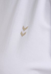 Witte katoenen t-shirt met een subtiel, goudkleurig chevron-embleem aan de onderste linkerkant. Glad textuur, eenvoudig ontwerp.