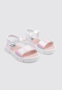 Sandalias con una base blanca, que presentan una correa rosa iridiscente brillante. Cierre de velcro, suela plana y diseño de plantilla acolchada.