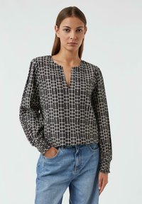 Schwarz-weißt gemusterte Bluse mit tiefem V-Ausschnitt, langen Ärmeln und elastischen Manschetten, kombiniert mit hellblauen Jeans. Strukturiertes Material.