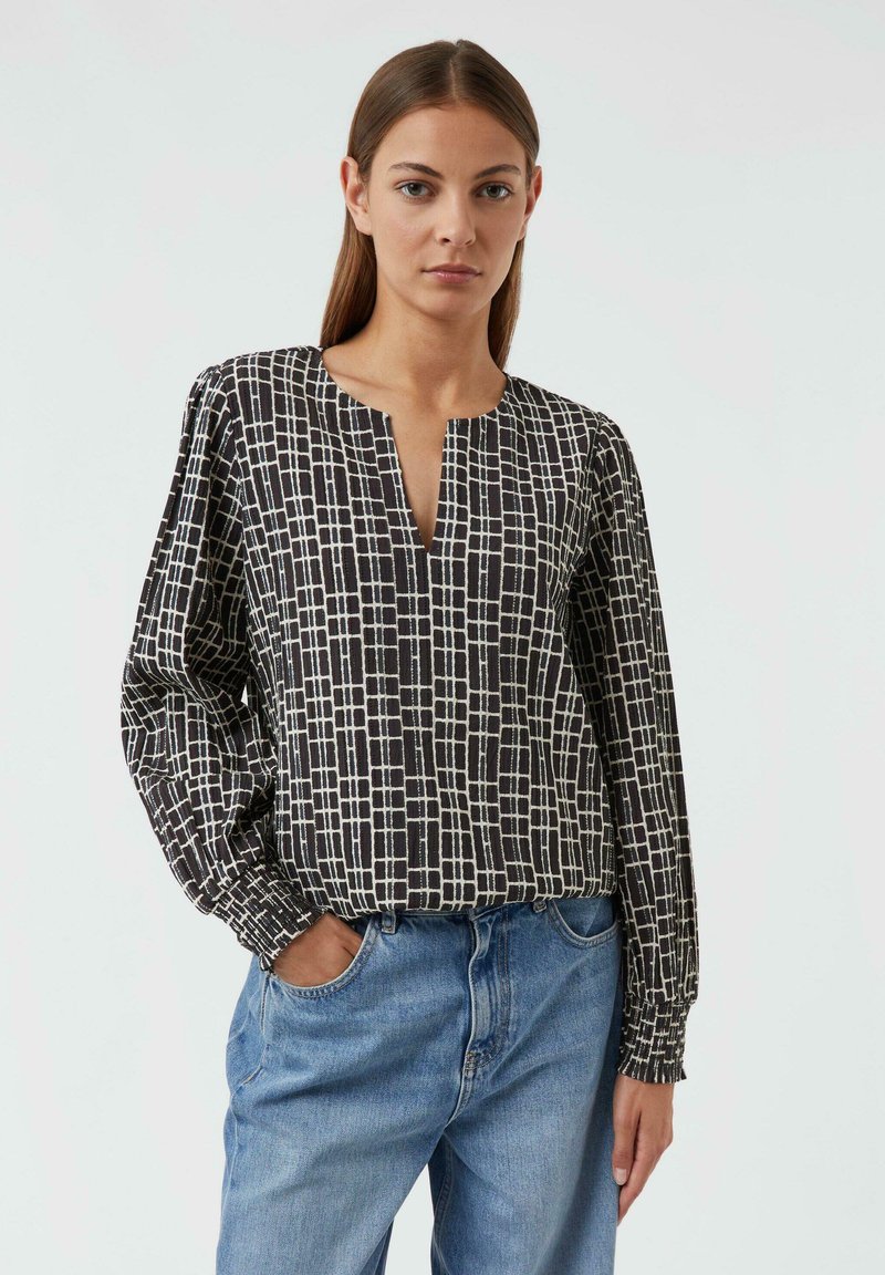 Schwarz-weißt gemusterte Bluse mit tiefem V-Ausschnitt, langen Ärmeln und elastischen Manschetten, kombiniert mit hellblauen Jeans. Strukturiertes Material.