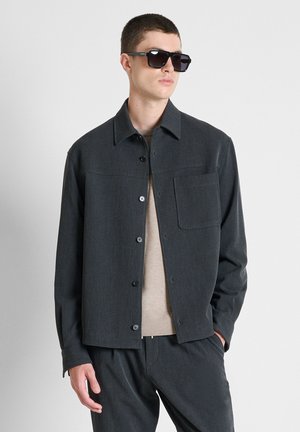 Man draagt donkergrijze jas met knopen, bijpassende broek, beige trui en zwarte zonnebril, staand met één hand in de zak.