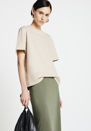 Maglietta beige a maniche corte con vestibilità rilassata, abbinata a una lunga gonna verde oliva. La modella tiene in mano una borsa nera con texture.