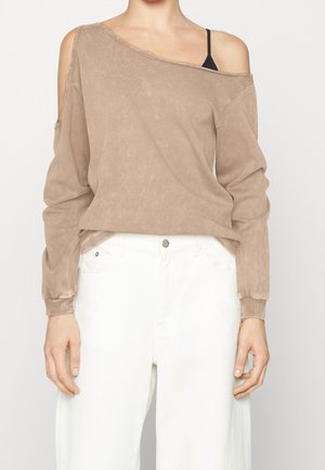Personne portant un haut beige à épaules dénudées avec manches longues, une bretelle noire visible et un pantalon blanc taille haute sur un fond uni.