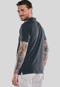 Dunkelgraues kurzärmeliges Polohemd mit einer lockeren Passform, seitlichen Schlitzen und subtilen Stickdetails, getragen über gestreiften Hosen.