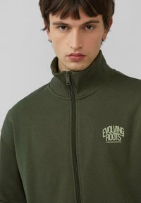 Khakigrüner Reißverschluss-Pullover mit hohem Kragen, der einen dezenten Logo-Druck auf der linken Brust und eine Rippstruktur aufweist.