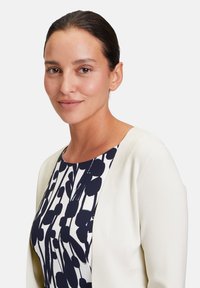 White cardigan over a navy and white floral-patterned dress. The fabric is smooth with a rounded neckline and short sleeves, creating a layered look.