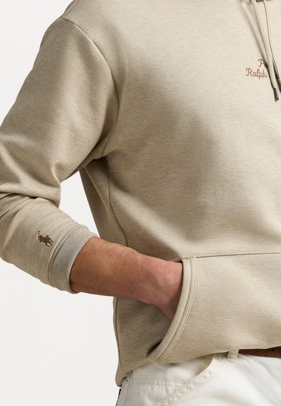 Beige hoodie med en frontlomme, ribbede manchetter og snørebånd. Har broderet logo på brystet og et lille emblem på ærmet.