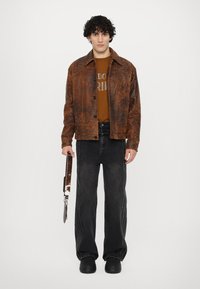 Giacca marrone con texture e due tasche frontali, t-shirt grafica nera sotto, jeans neri a gamba larga e scarpe nere. Sta tenendo una tracolla con un manico.