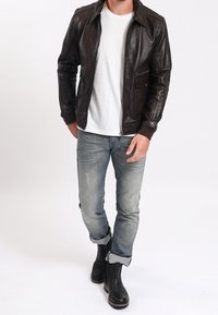 Hnědá kožená bunda se zipovým zapínáním, dvě přední kapsy, žebrované manžety, nošená s bílým tričkem a světle modrými džínami. Černé boty.