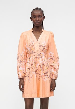 Femme aux cheveux tressés portant une robe longue à fleurs pêche, manches longues, col en V et taille froncée, debout devant un fond uni.