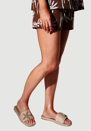 Frau trägt braune Shorts mit weißen Blattmustern und beige gewebte Slide-Sandalen mit einem Perlen-Sternfisch-Design, steht mit gekreuzten Beinen.